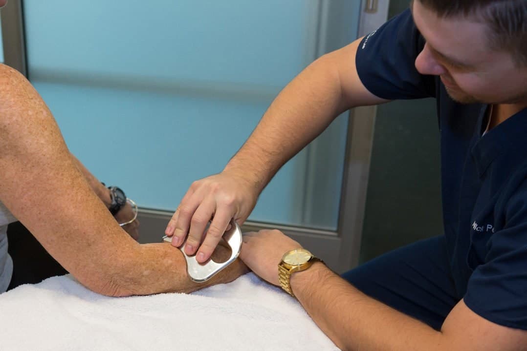 A therapist using a stainless steel instrument on a patient's elbow