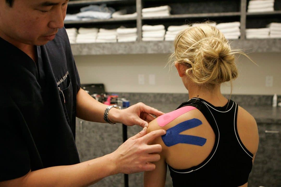 Davis Koh, DPT, applying pink and blue kinesiology tape to the back of a patient's shoulder