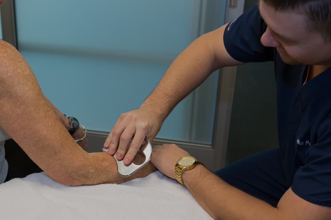 A KOH Physical Therapy Lab therapist using a stainless steel tool for soft tissue work on a client's forearm and elbow