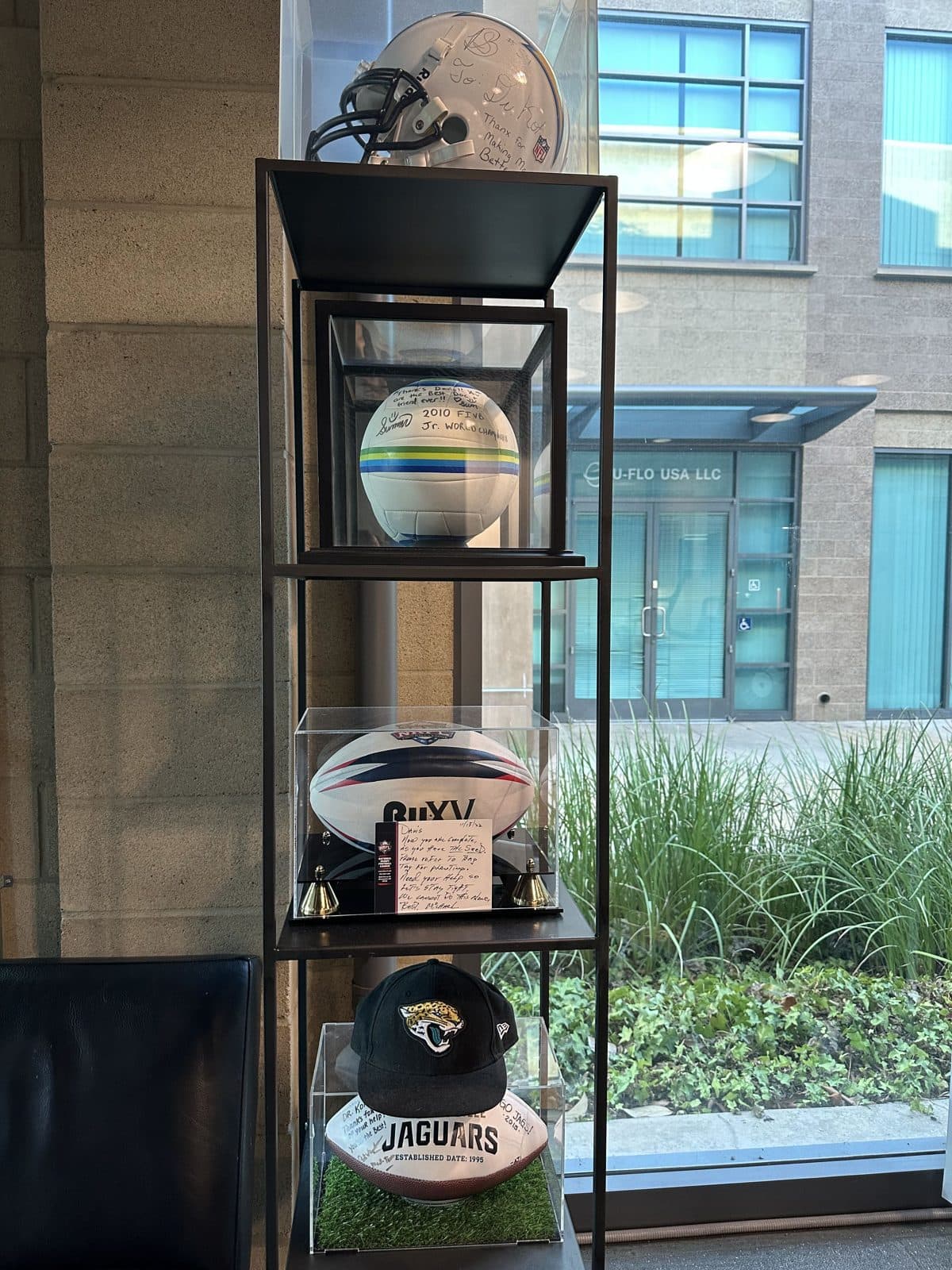 A display shelf by the window holding a signed helmet, volleyball, rugby ball and football