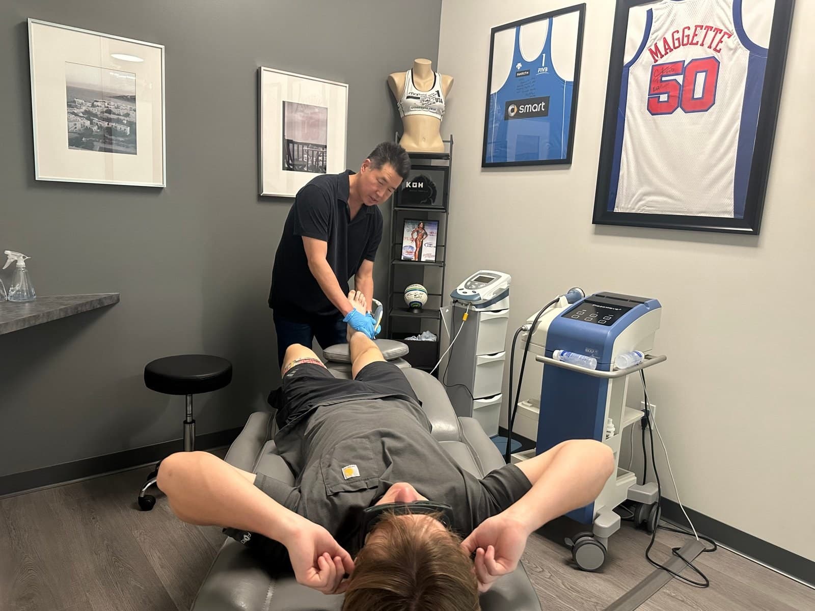 Davis Koh, DPT, treating a patient's foot with the shockwave applicator in a treatment room with signed athlete jerseys on the wall